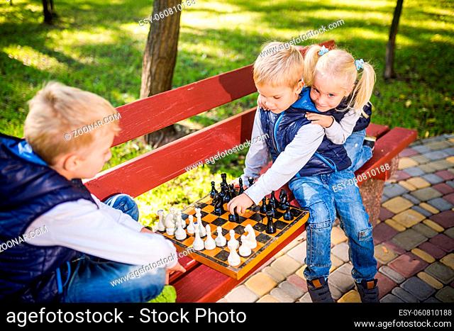 Little schoolchildren playing chess in the park on the bench. Summer holidays, intellectual games. Chess club for kids. Happy childhood and kids friendship