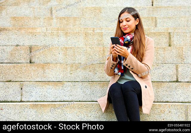 Beautiful stylish woman sitting on street stairs using mobile phone on winter day
