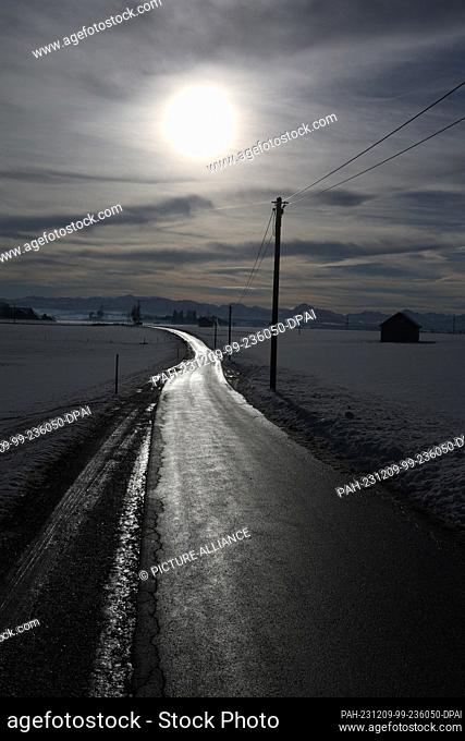 09 December 2023, Bavaria, Ruderatshofen: A road in a wintry landscape glows in the sunshine against the panorama of the Alps