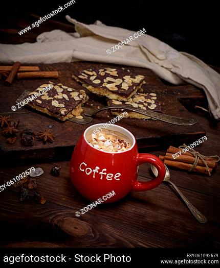 red ceramic cup with coffee and pieces of white marshmallow on a brown wooden background, behind chocolate baked with almonds