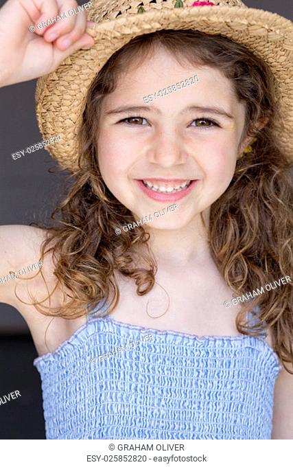 Portrait of a young girl with a grey background. She is wearing a blue dress and a straw hat and smiling at the camera