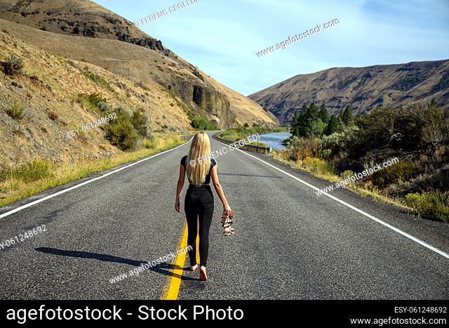 Attractive blond woman holding her shoes in her hand and walking away from it all on an empty countryside road