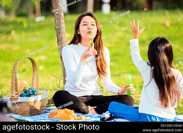 Happy Asian mother and little girl daughter child having fun and enjoying outdoor together sitting on the grass blowing soap bubbles during a picnic in the...