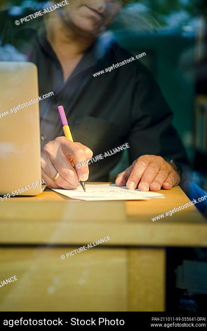 PRODUCTION - 01 October 2021, Bavaria, Munich: A man is sitting at a dining table at home. He is writing with a pen on a piece of paper next to a laptop