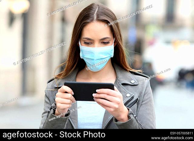 Serious woman with protective mask avoiding contagion watching video online on smart phone in the street