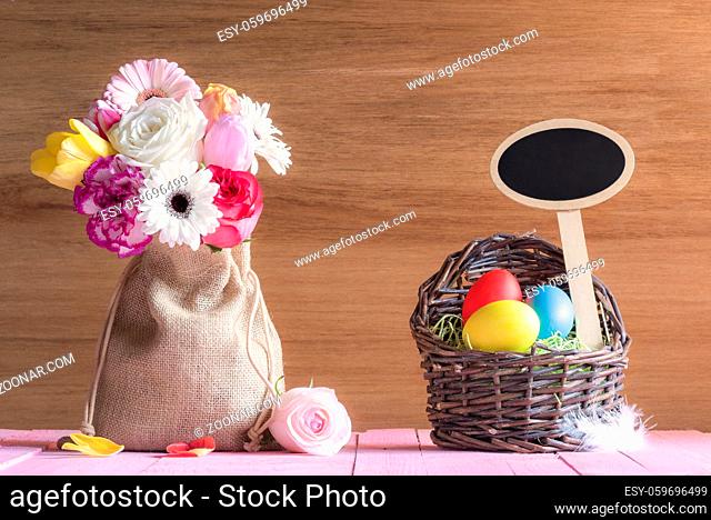 Bouquet of flowers in a jute sack and a wicker basket with painted eggs and a blank wooden banner, on a pink table, with a wooden wall