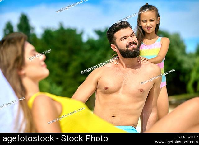 Young female dozing in the sunlounger outdoors while her smiling husband and cute daughter sunbathing