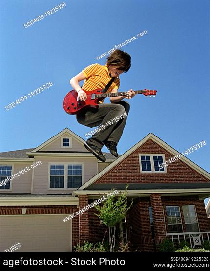 Teenage boy with guitar jumping in front of home, striking a pose mid air