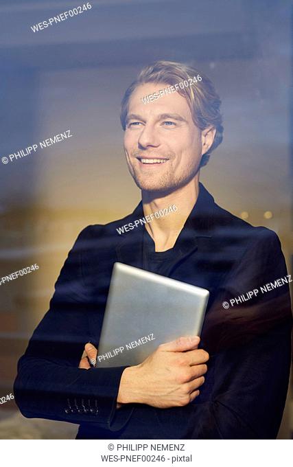 Portrait of smiling young businessman with tablet behind windowpane