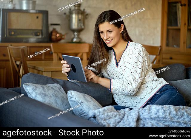 beautiful young smiling woman in white sweater sitting with a tablet on a gray sofa