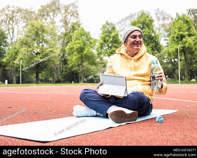 Smiling senior man with tablet PC and water bottle sitting on exercise mat