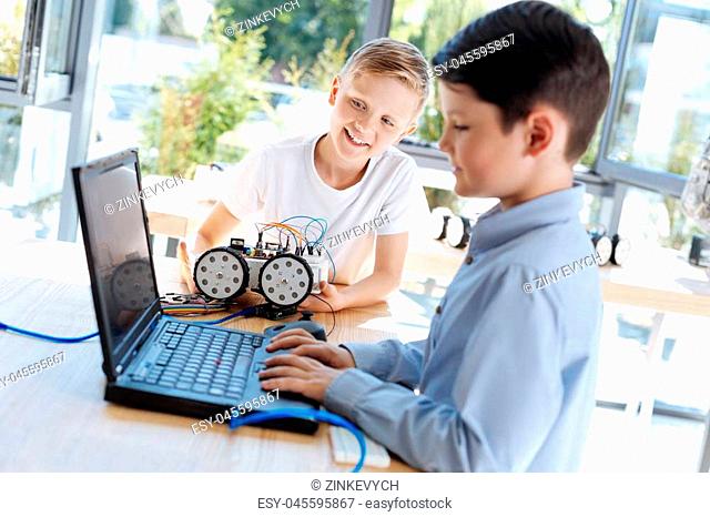 Little genius. Handsome dark-haired boy searching something via the laptop during the robotics workshop while his friend holding a car model and looking at...