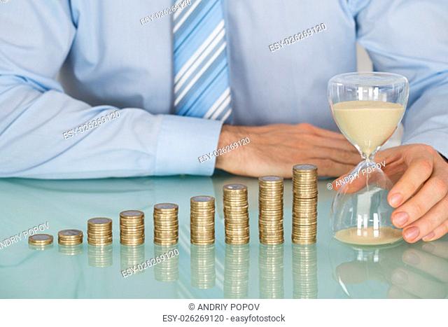 Close-up Of Businessman Holding Hourglass Near Stack Of Coins