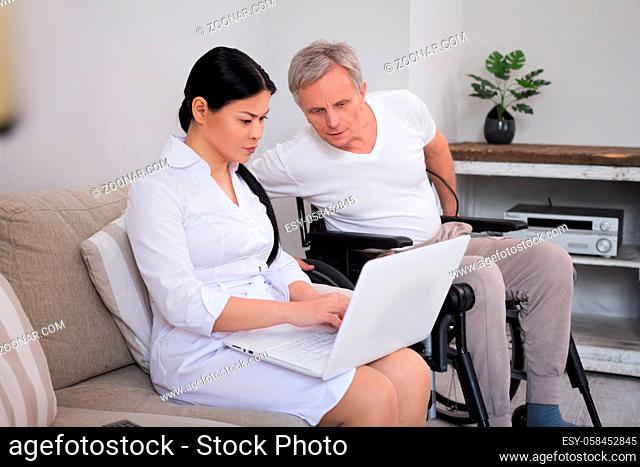 Nurse typing on laptop sitting next to man in wheelchair. Pretty female doctor sitting on couch showing her elderly male disbaled patient something on computer