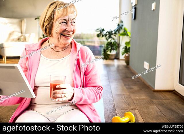 Cheerful senior woman with glass of juice sitting on ground at home