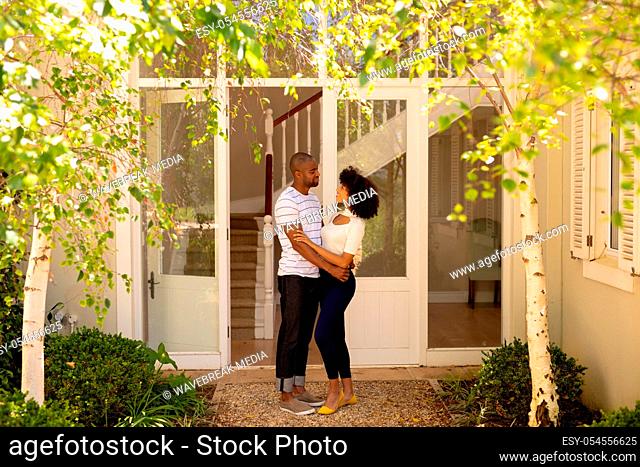 Side view of a mixed race couple standing in the garden outside their house on a sunny day, embracing and smiling at each other