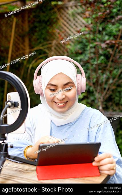 Smiling female influencer wearing headphones using tablet while vlogging at cafe