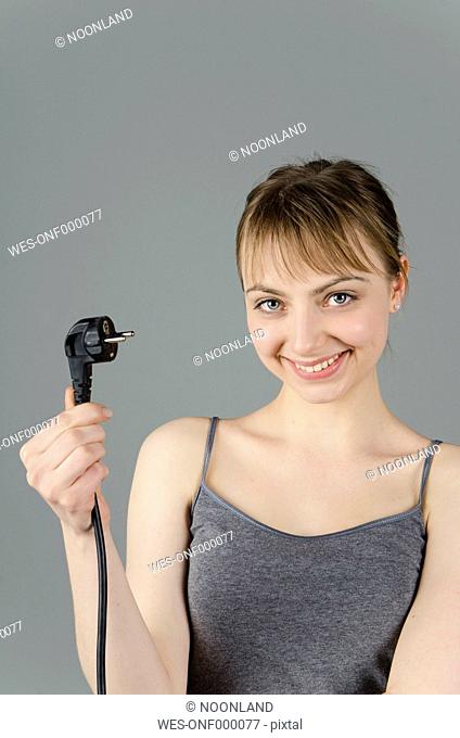 Portrait of young woman holding power plug against grey background