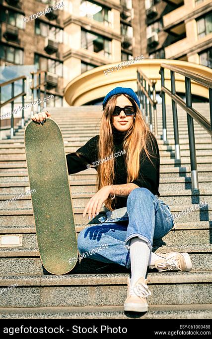 Young caucasian woman posing on street with skateboard in hands. Teenager girl in blue jeans extreme sports in an urban environment