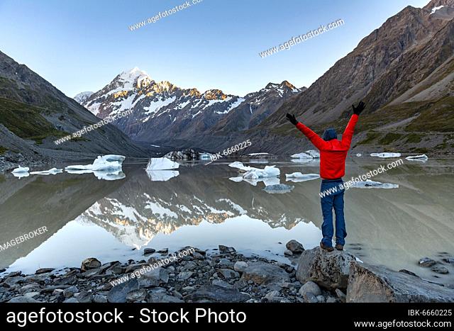 Hiker, young man standing on a rock and stretching his arms in the air, Mount Cook in morning light, Hooker Lake with reflection, sunrise