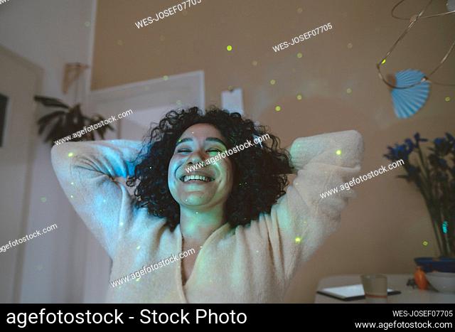 Happy woman with hands behind head and eyes closed sitting under astro lights at home