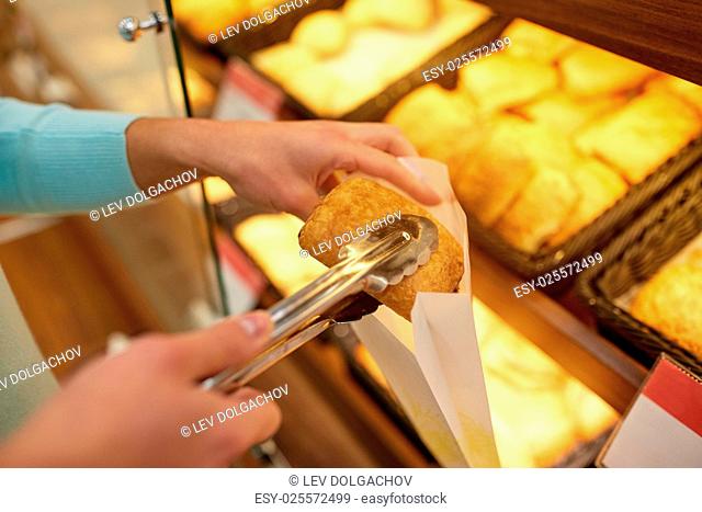 food, baking and sale concept - close up of hand with tongs and paper bag buying bun at bakery or grocery store