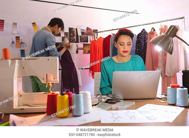 Female fashion designer working on laptop while male fashion designer dressing mannequin