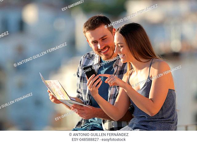 Happy couple of tourists comparing smartphone content and paper map in a town on vacation