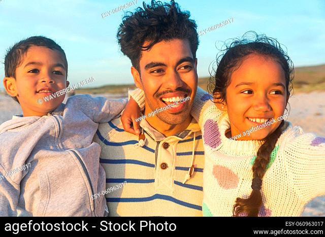 Happy biracial father carrying son and daughter looking away at beach during sunset