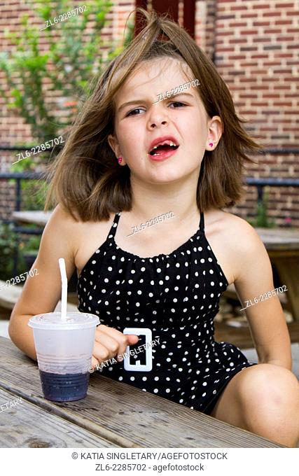 Caucasian young 8 years old, making a face. She has no front teeth and wear a polka dotted black and white dress. She is sitting at a wood table and sipping a...