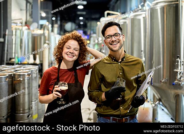Portrait of happy man and woman holding clipboard and beer glass in craft brewery