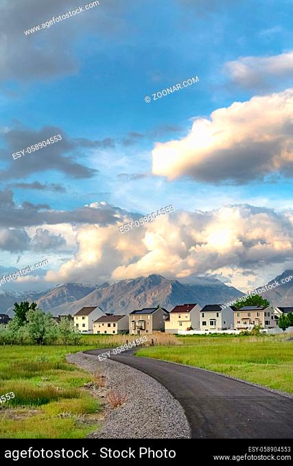 Paved road that curves through a field with multi storey homes in the distance. Huge mountain and cloudy blue sky can also be seen in this scenic landscape