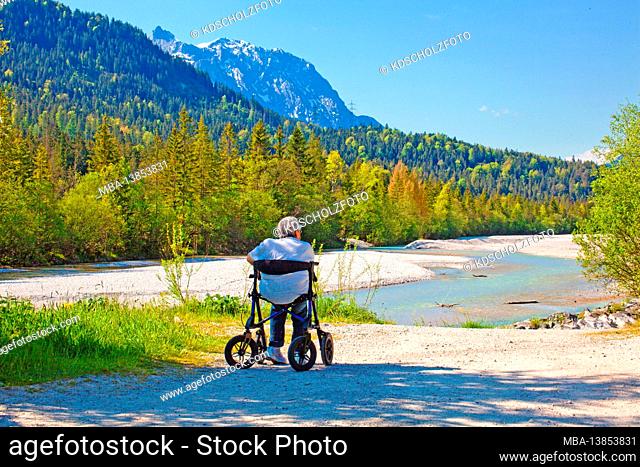 Old woman sits in a rollator on the banks of the Isar near Wallgau and enjoys the beautiful landscape