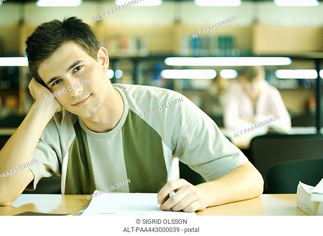 Male college student sitting in library, smiling at camera