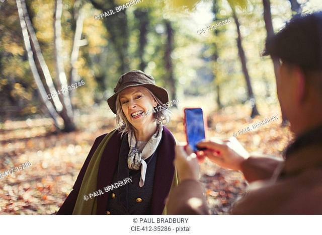 Playful senior woman being photographed by husband with camera phone in autumn park