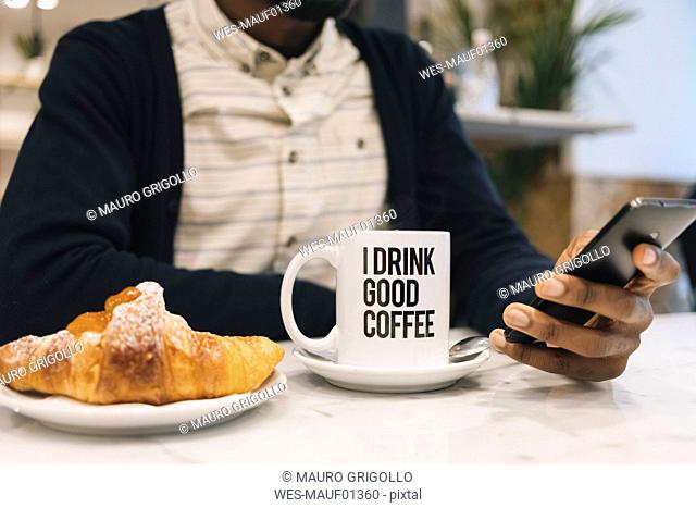 Close-up of man with croissant and cup of coffee in a cafe using cell phone