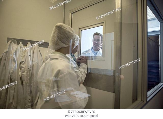 Two lab technicians in sterile protective clothing looking at colleague behind glass pane