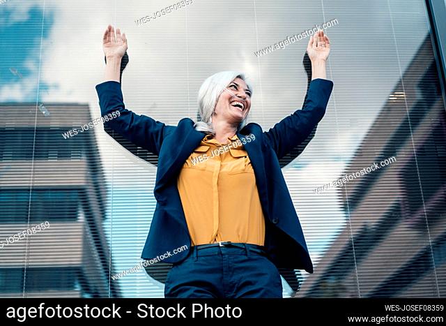Cheerful businesswoman with arms raised in front of glass wall