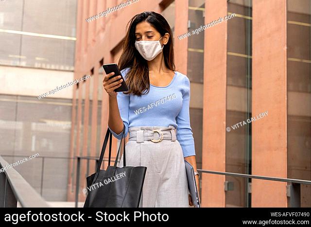 Businesswoman in face mask using smart phone while walking against office building during pandemic