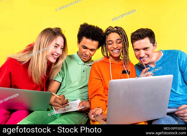 Smiling friends studying together on laptop in front of yellow background