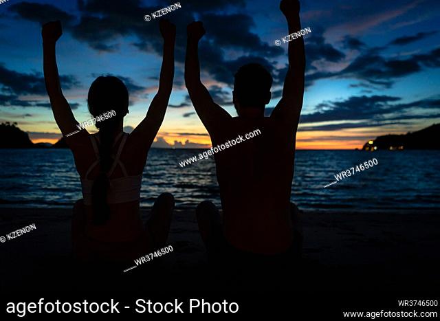 Rear view of a young couple sitting on the beach with raised arms while feeling happy and free during summer vacation in Flores Island, Indonesia