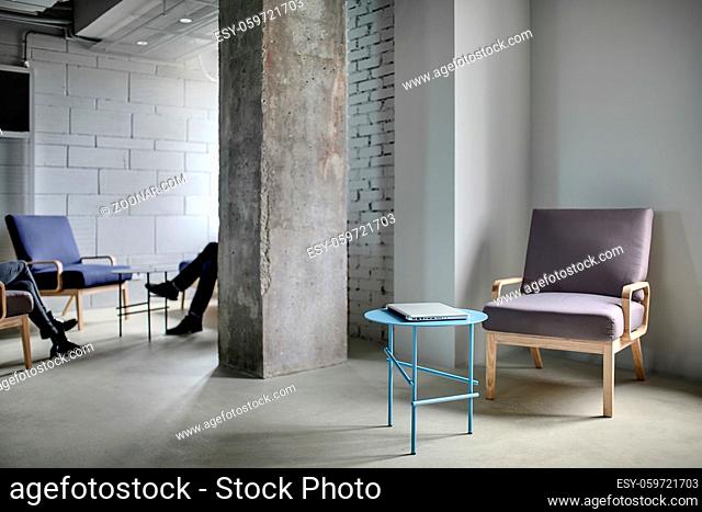 Zone in the office in a loft style with a concrete column and light walls. Near the column there are multi-colored armchairs with small round tables with a...