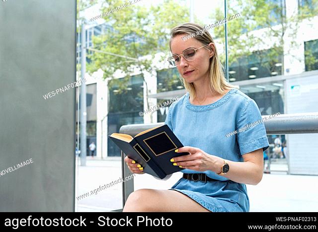 Blond businesswoman reading book while sitting in front of glass wall