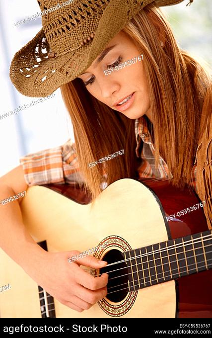 Attractive young woman playing guitar with joy, wearing western hat