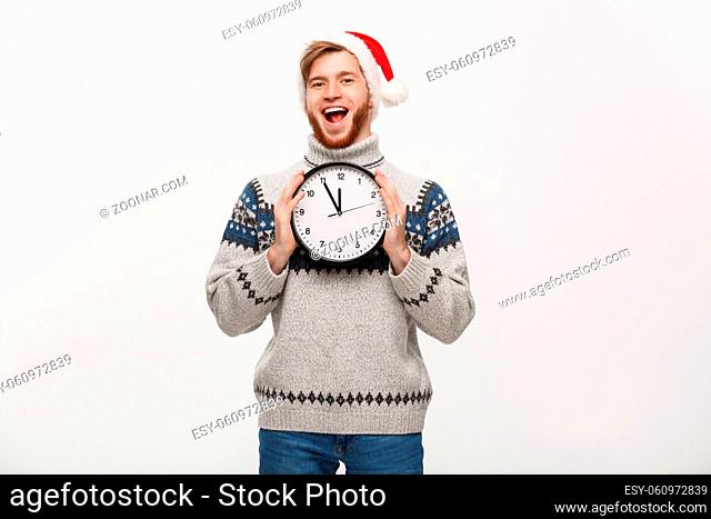 Holiday concept - Young happy handsome beard man in sweater holding white clock over white studio background
