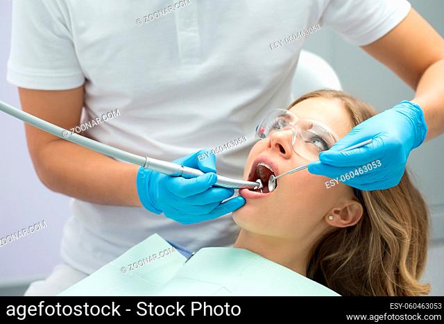Brave girl with opened mouth in patient bib and protective eyewear. Next to her there is a dentist in a white uniform with blue latex gloves