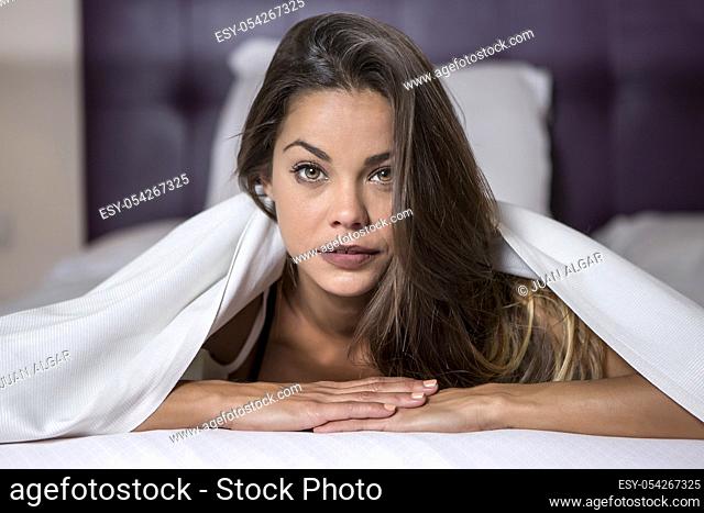 Young charming woman with long silky hair lying under white blanket on bed and looking at camera