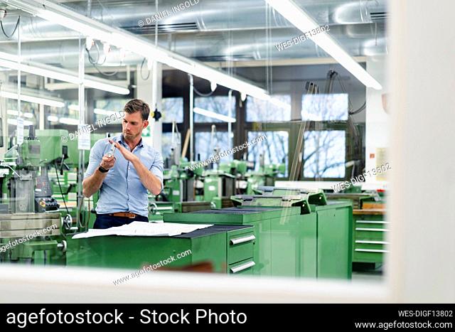 Male professional with blueprint checking machine part seen through glass in factory