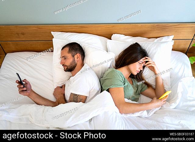 Young couple using smart phones while lying back to back in bed at home