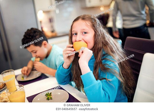 Little girl is looking at the camera while eating a slice of orange at home with her brother